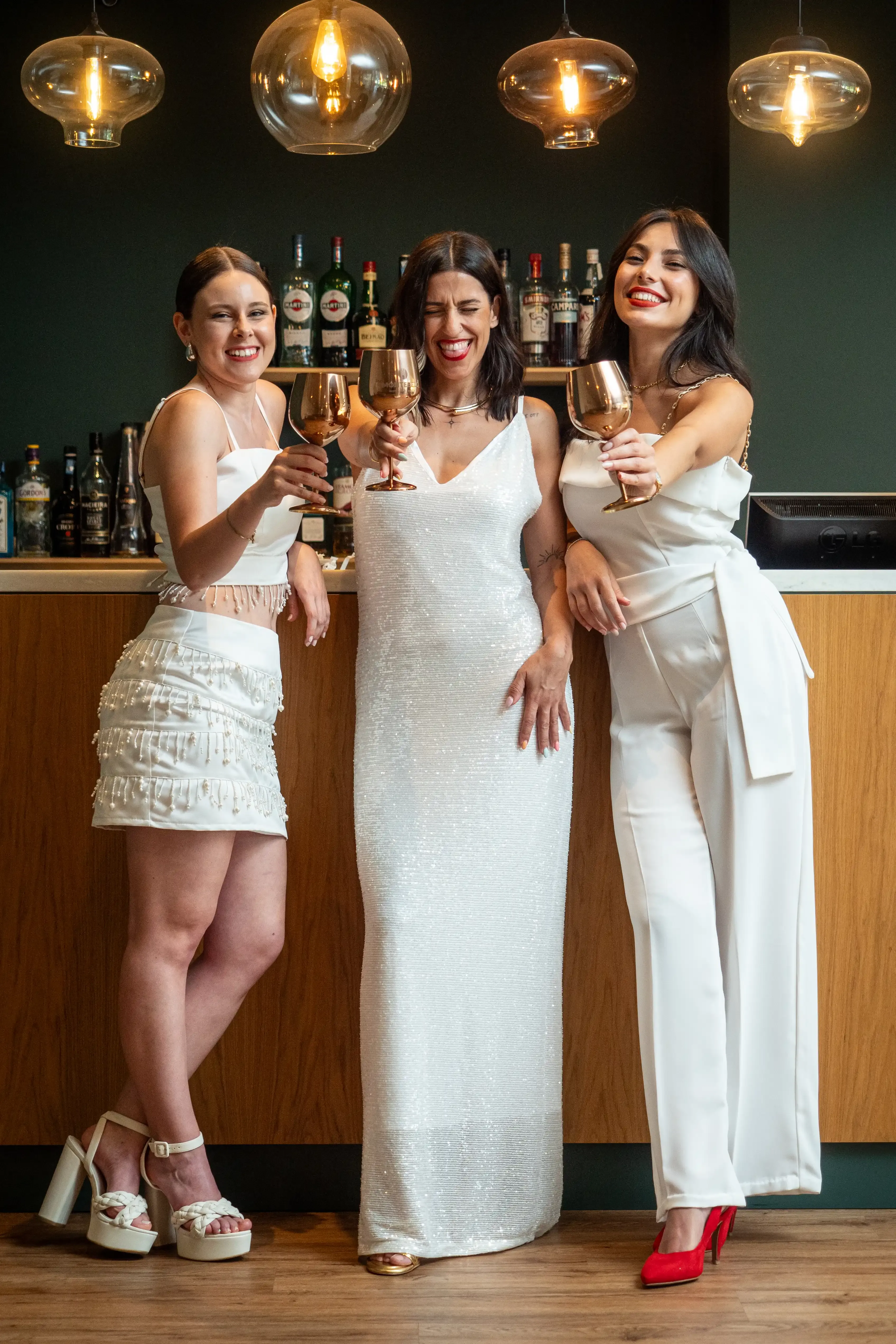 Três mulheres estão a posar enquanto brindam para a câmara. Estão vestidas de branco.